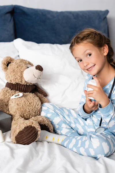 Smiling child holding pills bottle while with teddy bear in bedroom — Stock Photo