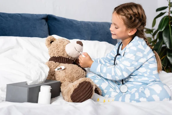 Niño curando oso de peluche con aerosol nasal mientras juega en la cama - foto de stock