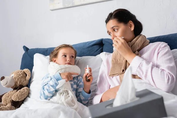 Больная девочка указывает на носовой спрей рядом с матерью, вытирающей нос бумажной салфеткой — стоковое фото