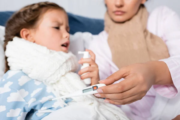 Donna malata che tiene il termometro mentre la figlia utilizza lo spray alla gola su sfondo sfocato — Foto stock