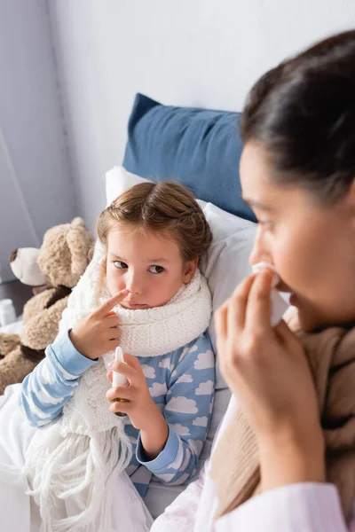 Menina doente segurando spray nasal perto da mãe sofrendo de corrimento nasal em primeiro plano borrado — Fotografia de Stock
