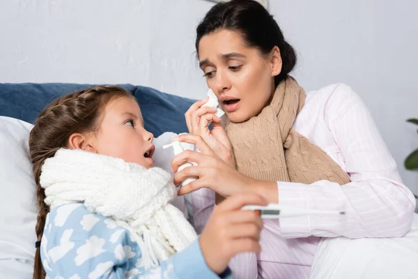 Femme malade avec nez qui coule tenant pulvérisation de gorge près de fille malade avec thermomètre sur le premier plan flou — Photo de stock