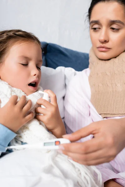 Upset mother holding thermometer while sick daughter using throat spray — Stock Photo