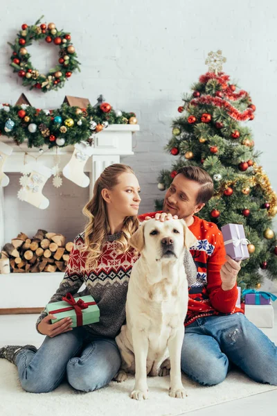 Joyful couple holding presents and sitting near dog and christmas tree — Stock Photo