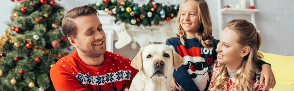 Família feliz em suéteres olhando para labrador perto de árvore de natal decorada, banner — Fotografia de Stock