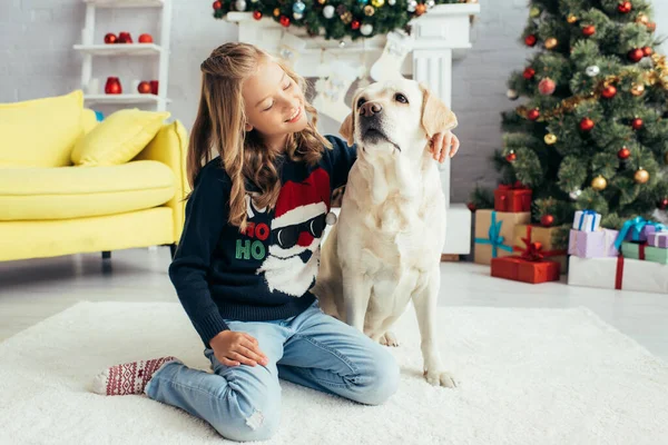 Chica feliz en suéter caliente sentado cerca de labrador en sala de estar decorada, concepto de Navidad - foto de stock