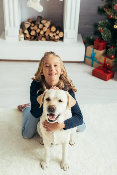 Menina feliz em camisola quente abraçando labrador na sala de estar decorada, conceito de Natal — Fotografia de Stock