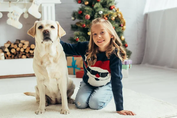 Fröhliches Kind im warmen Pullover sitzt neben Labrador im geschmückten Wohnzimmer, Weihnachtskonzept — Stockfoto