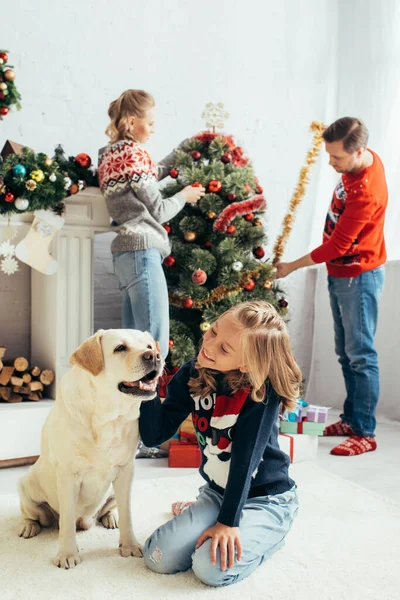Selective focus of joyful girl in sweater cuddling labrador near parents decorating christmas tree in living room — Stock Photo