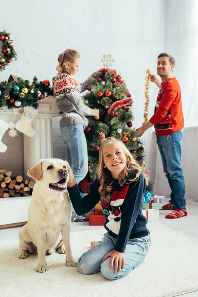 Selective focus of joyful girl in sweater cuddling labrador near happy parents decorating christmas tree in living room — Stock Photo