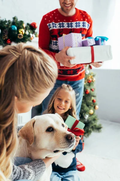 Mujer abrazando perro cerca de hija y marido con regalos en Navidad - foto de stock