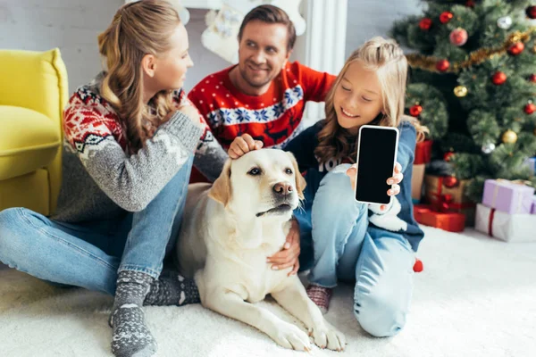 Niño alegre abrazo perro mientras sostiene el teléfono inteligente con pantalla en blanco cerca de los padres en la sala de estar decorada - foto de stock