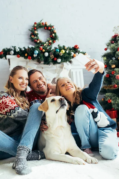 Niño tomando selfie con padres alegres y perro en sala de estar decorada - foto de stock