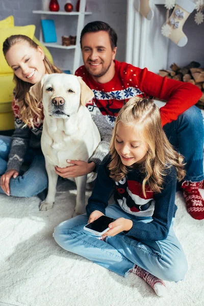 Bambino gioioso utilizzando smartphone con schermo bianco vicino ai genitori e cane a casa — Foto stock
