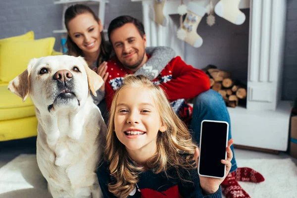 Niña sosteniendo teléfono inteligente con pantalla en blanco cerca del perro y los padres sobre fondo borroso - foto de stock