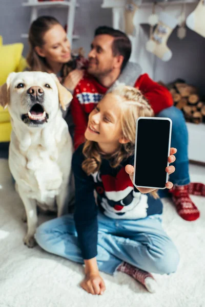 Heureuse fille tenant smartphone avec écran vierge et famille floue sur fond — Photo de stock