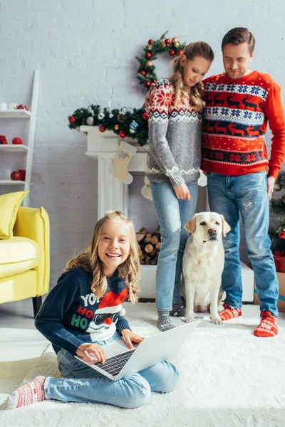 Happy kid using laptop near joyful parents standing near dog on christmas — Stock Photo