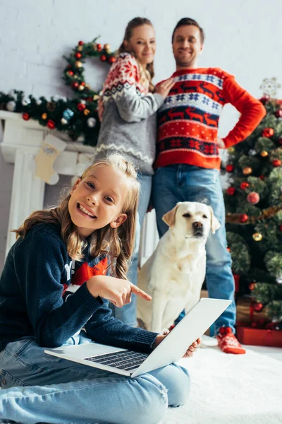 Happy kid pointing with finger at laptop near joyful parents standing near dog and christmas tree — Stock Photo