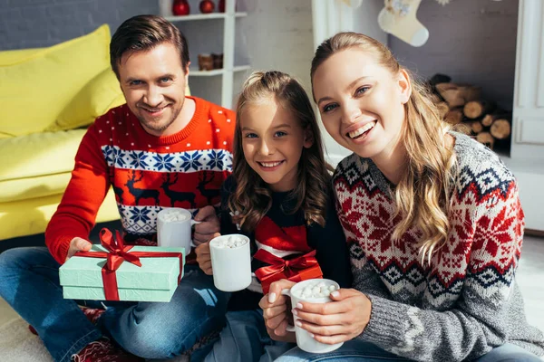 Familia en suéteres sosteniendo copas con bebidas cerca presente en Navidad - foto de stock
