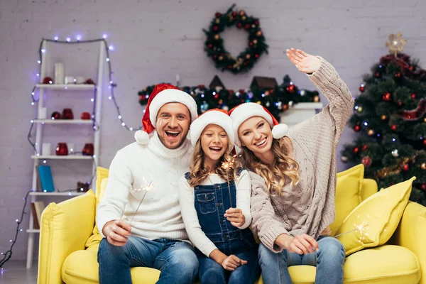 Família alegre em chapéus santa segurando sparklers na sala de estar decorada — Fotografia de Stock