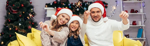 Щаслива сім'я в капелюхах Санти тримає блискітки в декорованій вітальні, банер — стокове фото