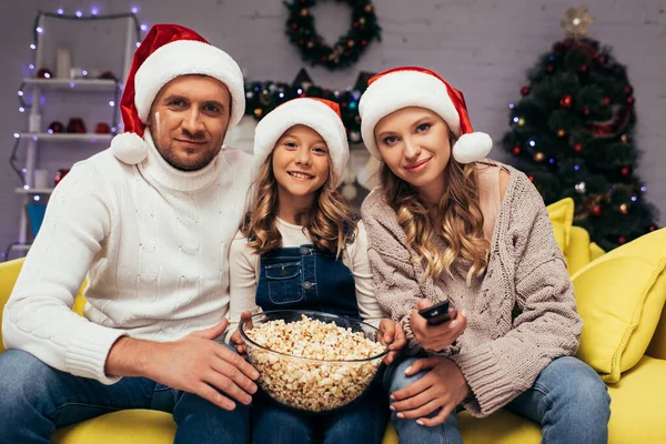 Glückliche Familie in Weihnachtsmützen blickt in die Kamera neben Schüssel mit Popcorn — Stockfoto