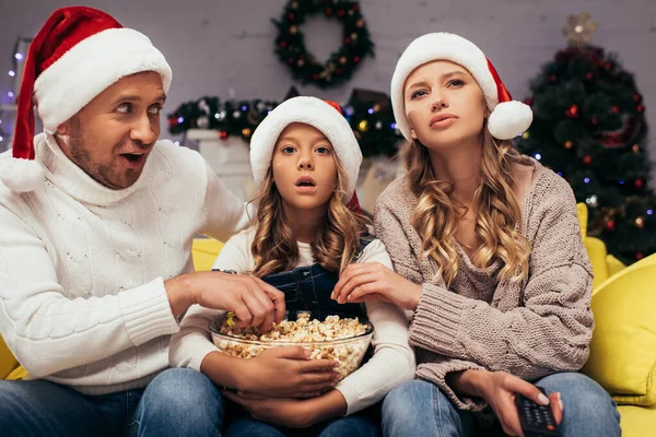 Famiglia scioccato in cappelli di Babbo Natale guardando la fotocamera vicino ciotola con popcorn — Foto stock