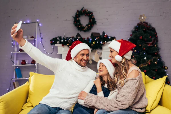 Familia alegre en sombreros de santa tomar selfie en sala de estar decorada - foto de stock