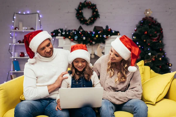 Joyful kid in santa hat using laptop near parents in decorated living room — Stock Photo