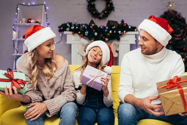 Freudige Eltern mit Weihnachtsmützen, die Geschenke in der Hand halten und das Kind an Weihnachten im geschmückten Wohnzimmer anschauen — Stockfoto
