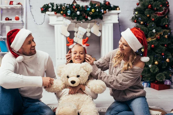Mother wearing headband with reindeer horns on daughter with teddy bear near husband — Stock Photo