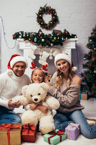 Niño feliz en diadema con cuernos de reno sosteniendo oso de peluche cerca de regalos y padres en sombreros de santa - foto de stock