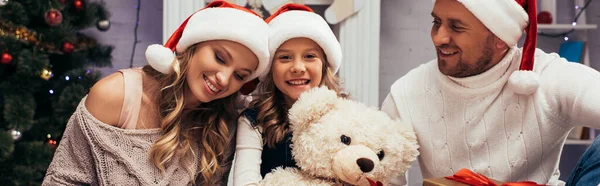 Счастливый ребенок держит плюшевого мишку возле подарков и родителей в шляпах Санта-Клауса, баннер — стоковое фото