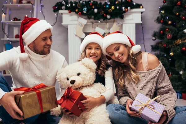 Bambino felice tenendo orsacchiotto vicino regali e genitori in cappelli di Babbo Natale — Foto stock