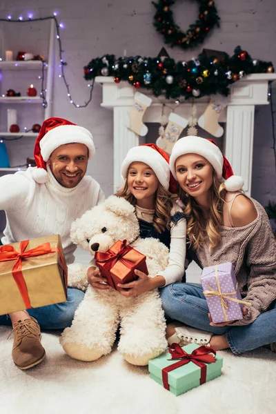 Família alegre em chapéus santa segurando presentes na sala de estar decorada no Natal — Fotografia de Stock