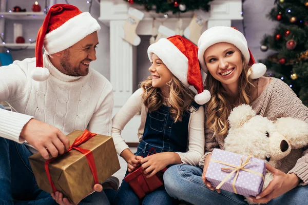 Família satisfeita em chapéus santa segurando presentes na sala de estar decorada no Natal — Fotografia de Stock