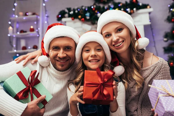 Glückliches Kind mit Weihnachtsmütze hält Weihnachtsgeschenk in der Nähe der Eltern — Stockfoto