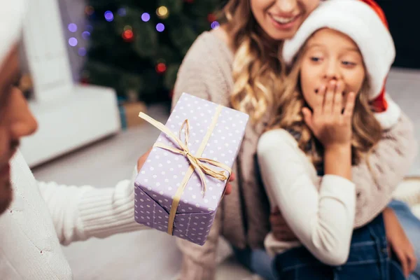 Heureux père dans santa chapeau donnant cadeau à la fille surprise avec femme floue sur fond — Photo de stock