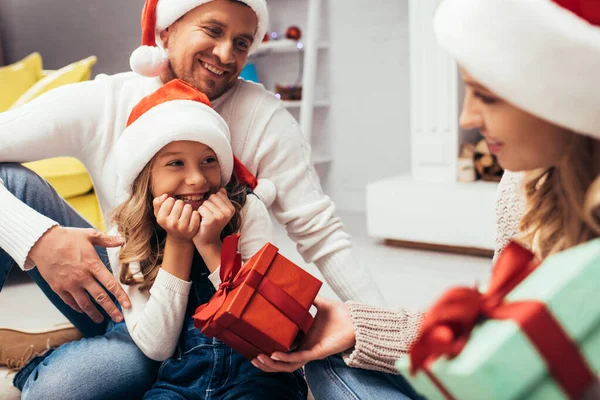 Mère dans santa chapeau donnant cadeau à fille heureuse près du père — Photo de stock