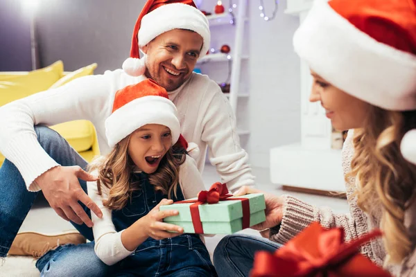 Мати в капелюсі Санта дарує подарунок збудженій дочці поблизу батька — стокове фото