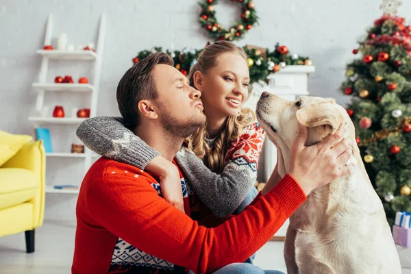 Happy man in knitted sweater cuddling labrador near wife in decorated apartment on christmas — Stock Photo