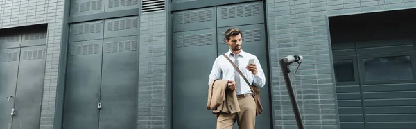 Geschäftsmann im Anzug hält Smartphone in der Nähe von E-Scooter und Gebäude, Banner — Stockfoto