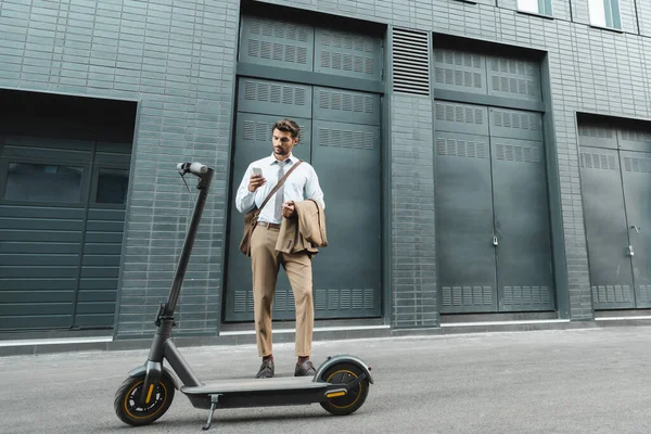 Full length of businessman in suit holding smartphone while standing near e-scooter and building — Stock Photo