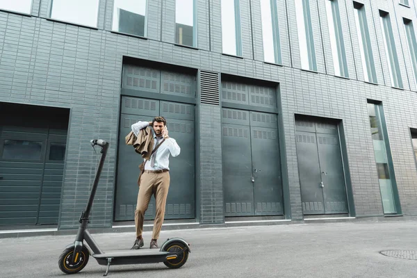 В полный рост бизнесмен разговаривает по смартфону, стоя возле е-скутера и здания — стоковое фото