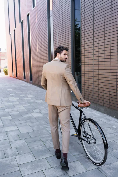 Full length of bearded businessman in suit walking with bike and looking away near building — Stock Photo