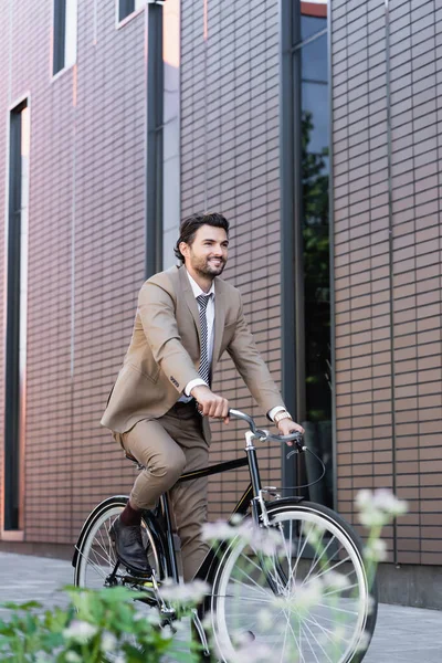 Felice uomo in giacca e cravatta in bicicletta e sorridente vicino edificio e impianto in primo piano sfocato — Foto stock