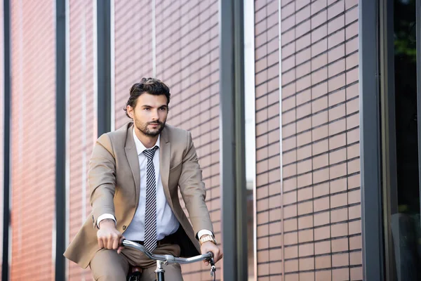 Hombre barbudo en traje de montar en bicicleta cerca de edificio moderno - foto de stock
