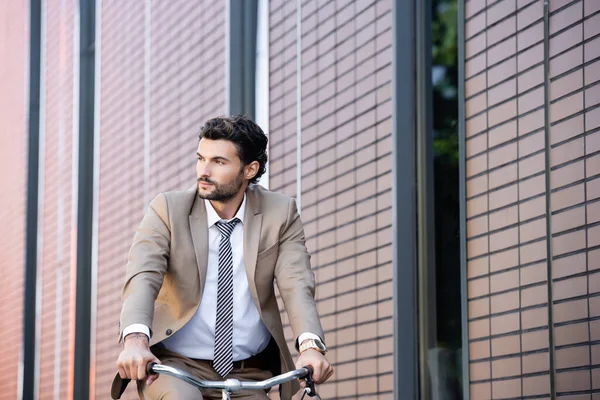 Homem de negócios barbudo em terno andar de bicicleta e olhando para longe perto do edifício — Fotografia de Stock