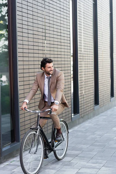 Full length of positive businessman in suit riding bicycle, smiling and looking away near building — Stock Photo