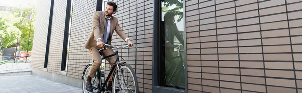 Homme d'affaires barbu en costume à vélo et regardant loin près du bâtiment, bannière — Photo de stock